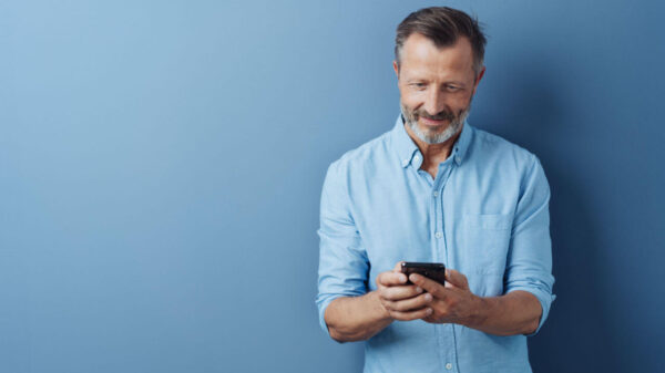 Homem de meia-idade lendo algo em seu celular, com um sorriso tranquilo, posando sobre um fundo de estúdio azul.