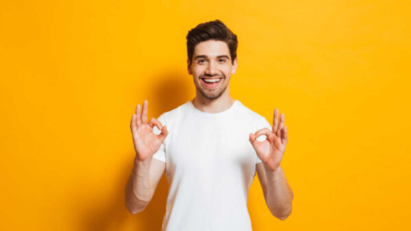 Retrato de homem animado, usando roupas básicas, sorrindo e fazendo sinal de "ok" para a câmera, isolado sobre fundo amarelo.
