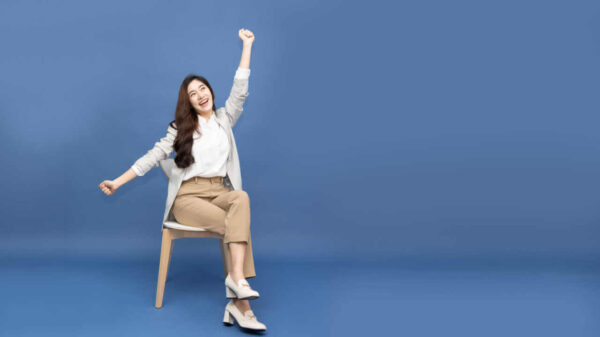 Mulher sentada em uma cadeira moderna e branca, com as mãos levantadas, com os braços erguidos de felicidade, isolada em um fundo azul. Conceito de sucesso.