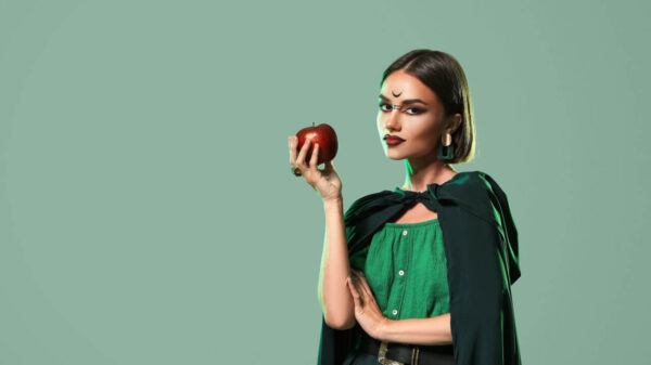 Mulher segurando uma maçã, vestida de bruxa, em fundo verde.