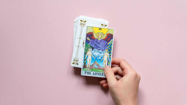 "Os Enamorados", carta de tarot Rider Waite, cima do baralho. Mão segurando a carta em fundo rosa.