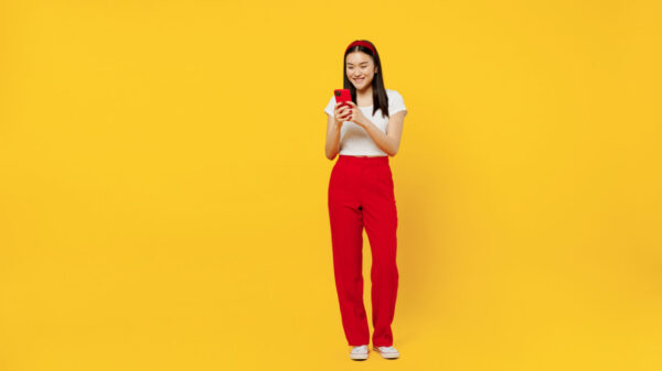 Comprimento do corpo em tamanho real de uma mulher jovem, sorridente, usando roupas casuais, usando um celular, isolada em fundo amarelo liso, em um retrato de estúdio.