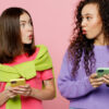 Duas amigas jovens chocadas, juntas, cada uma segurando um celular na mão, olhando uma para a outra, isoladas em um estúdio de fundo de cor rosa claro pastel.