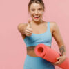 Mulher jovem fitness, atlética, forte, usando roupas azuis, mostrando o polegar para cima, piscando um olho, isolada em um fundo rosa claro pastel. Conceito de treino.
