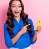 Foto de uma mulher animada e impressionada, apontando para o celular, isolada em um fundo de cor rosa.