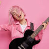 Foto de estúdio de uma musicista despreocupada, tocando uma música, mantendo o braço levantado, com cabelo rosa, segurando guitarra preta. Conceito de artista famosa.