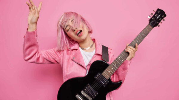 Foto de estúdio de uma musicista despreocupada, tocando uma música, mantendo o braço levantado, com cabelo rosa, segurando guitarra preta. Conceito de artista famosa.