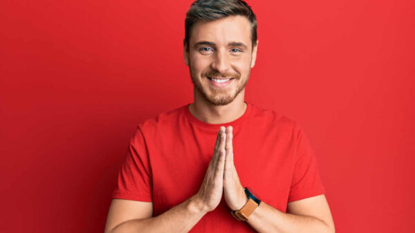 Homem vestindo camiseta vermelha casual, com as mãos juntas, sorrindo confiantemente.