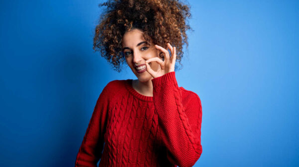 Mulher jovem, com cabelos cacheados e piercing, vestindo suéter vermelho casual, sorrindo positivamente, fazendo sinal de "ok" com a mão e os dedos. Expressão de sucesso.