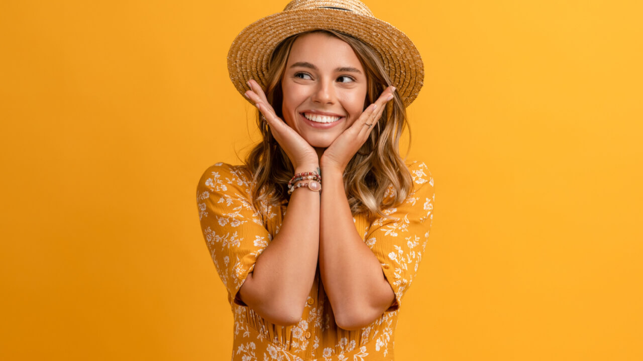 Mulher usando um vestido amarelo e um chapéu de palha, posando em um fundo amarelo isolado, com uma expressão de rosto sorridente e feliz.