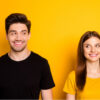 Foto de um casal alegre, sorridente e positivo. Duas pessoas, uma vestindo uma camiseta preta e a outra uma amarela. Os dois sorriem um para o outro, isolados sobre um fundo de cor amarela.