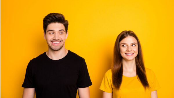 Foto de um casal alegre, sorridente e positivo. Duas pessoas, uma vestindo uma camiseta preta e a outra uma amarela. Os dois sorriem um para o outro, isolados sobre um fundo de cor amarela.