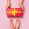 Vista superior de mãos segurando caixa de presente vermelha, com fita amarela, em um fundo rosa. Conceito de presente de, Dia dos Namorados.