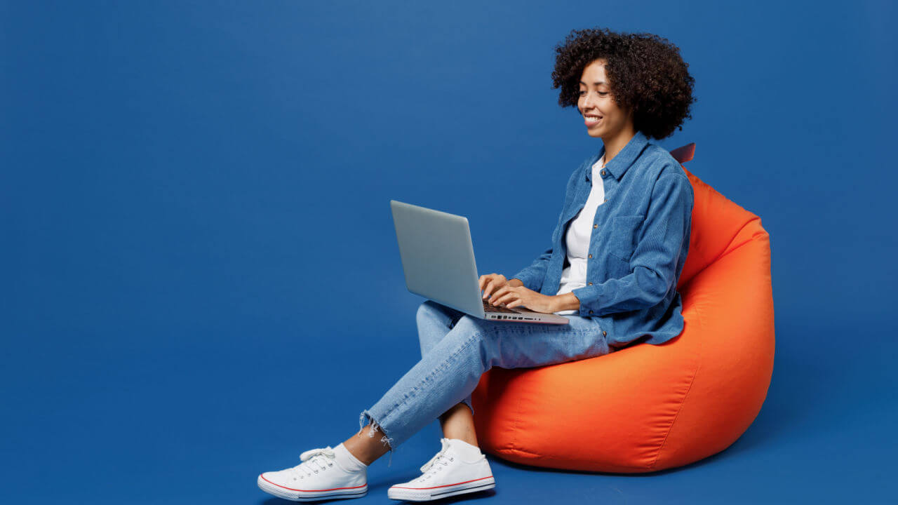 Mulher jovem sorrindo, em foto de corpo inteiro, feliz, usando roupas casuais, com uma camisa branca, sentada em um pufe, segure um computador, isolada em um estúdio de fundo azul escuro liso.