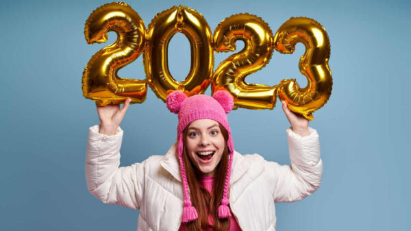Mulher jovem feliz, usando casaco de inverno, segurando números (2023) dourados, contra um fundo azul.