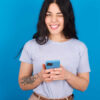 Mulher vestindo camiseta azul sobre fundo azul, piscando um olho, segurando um celular.