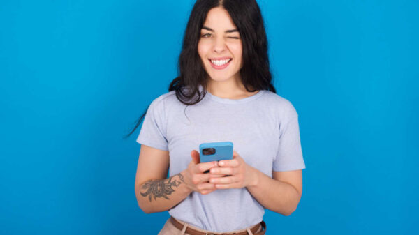 Mulher vestindo camiseta azul sobre fundo azul, piscando um olho, segurando um celular.