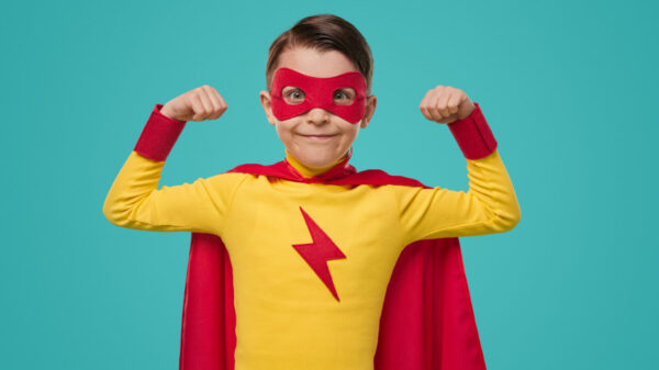 Garoto vestido de super-herói, com capa vermelha, blusa amarela e máscara vermelha, demonstrando poder, enquanto olha para a câmera, contra um fundo azul de um estúdio.