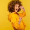 Retrato de uma mulher alegre, de cabelos cacheados, vestindo um casaco que tem um capuz, piscando um olho, apontando para a câmera, flertando. Foto de estúdio interior isolado em fundo amarelo.