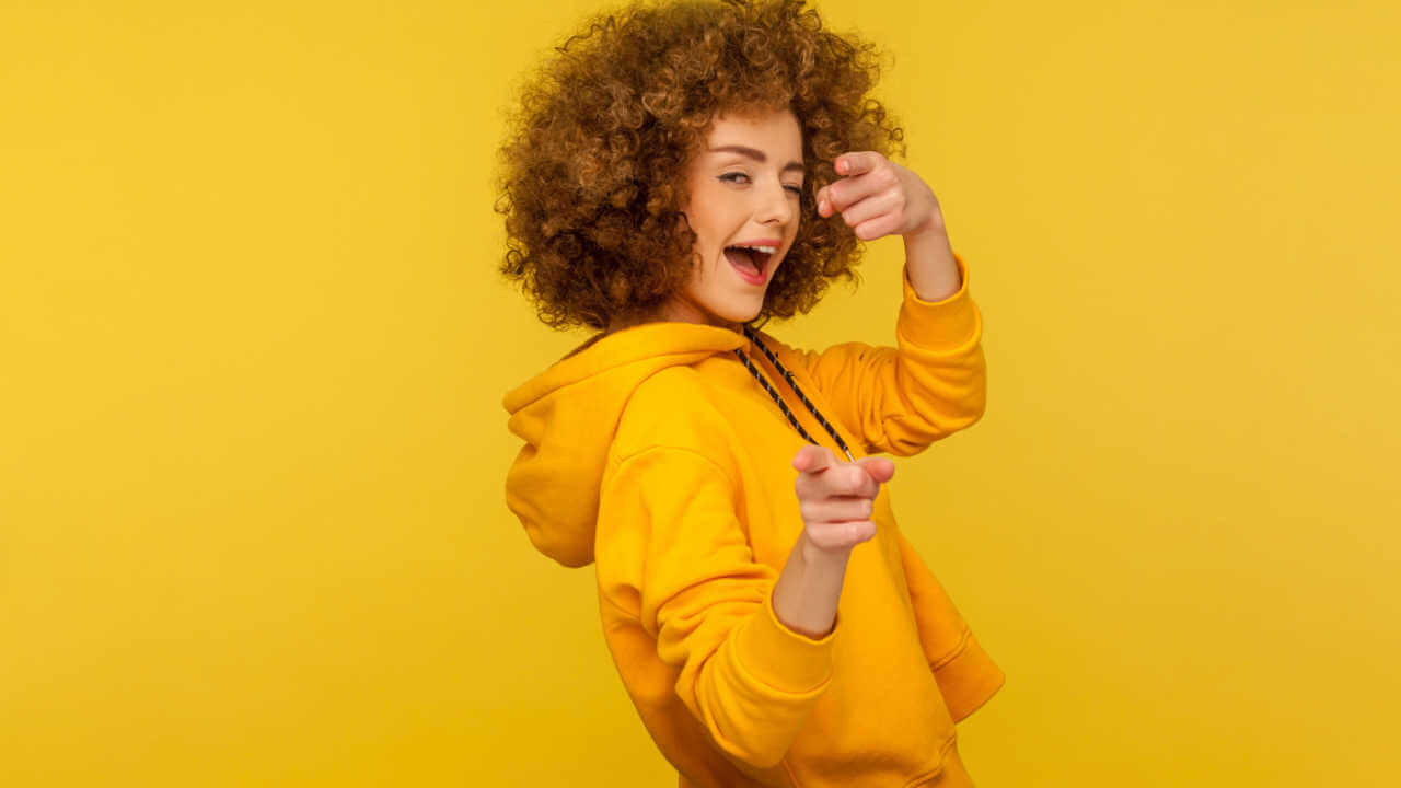 Retrato de uma mulher alegre, de cabelos cacheados, vestindo um casaco que tem um capuz, piscando um olho, apontando para a câmera, flertando. Foto de estúdio interior isolado em fundo amarelo.