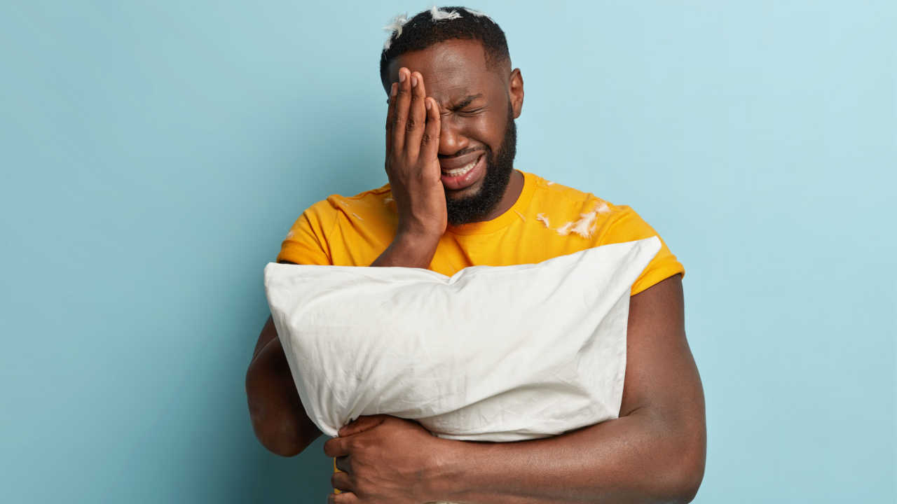 homem negro com roupa amarela e abraçando um travesseiro branco. sua mão está no rosto e está com expressão de emoção
