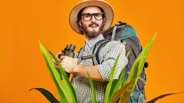 Conceito de turismo, viagens e aventura. Jovem com uma mochila nas costas e um chapéu de turista, espreitando por trás de plantas tropicais, segurando um binóculos. Retrato de estúdio em um fundo laranja.