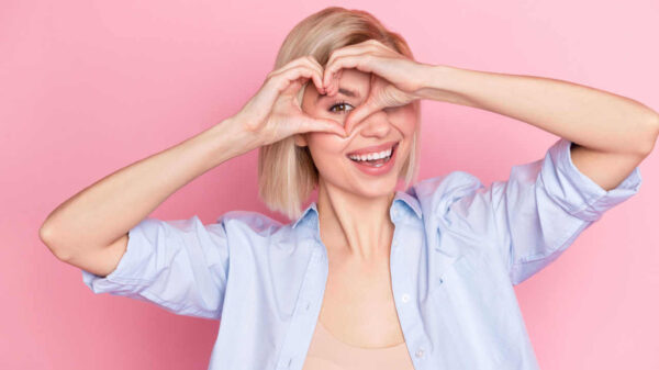 Foto de uma mulher e fazendo a forma de um coração com as mãoes na frente de um de seus olhos, isolada em um fundo de cor rosa pastel.