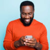 homem negro sorrindo olhando para o celular que está em suas mãos em fundo azul