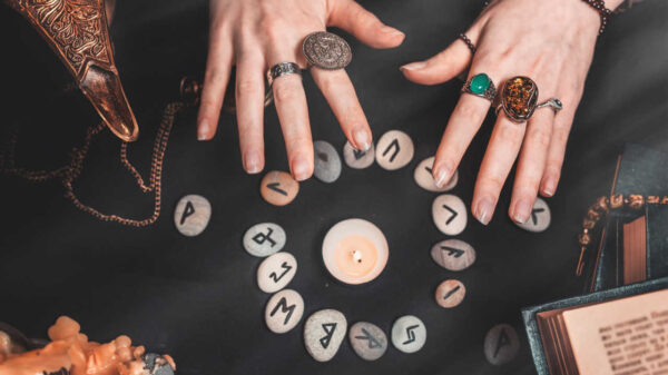 Astrologia e esoterismo. Mãos femininas sobre um círculo rúnico. Runas de adivinhação sobre um fundo preto, livros, amuletos preciosos, uma lâmpada de cobre e uma vela.