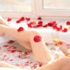 Mulher jovem deitada em uma banheira com espuma e com pétalas de rosas vermelhas, segurando um copo de vinho branco.
