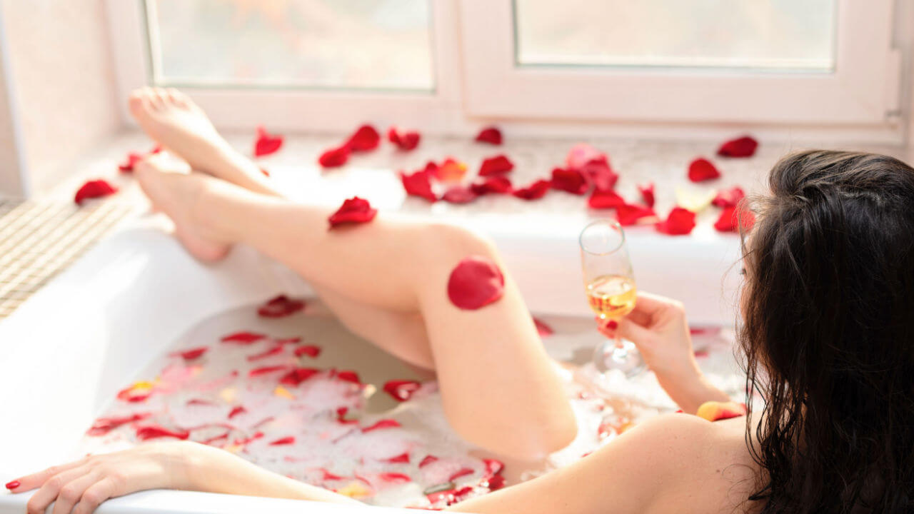 Mulher jovem deitada em uma banheira com espuma e com pétalas de rosas vermelhas, segurando um copo de vinho branco.