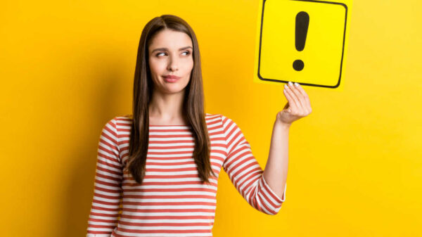 Retrato de uma garota duvidosa segurando um cartão com um ponto de exclamação, isolada sobre um fundo de cor amarela.