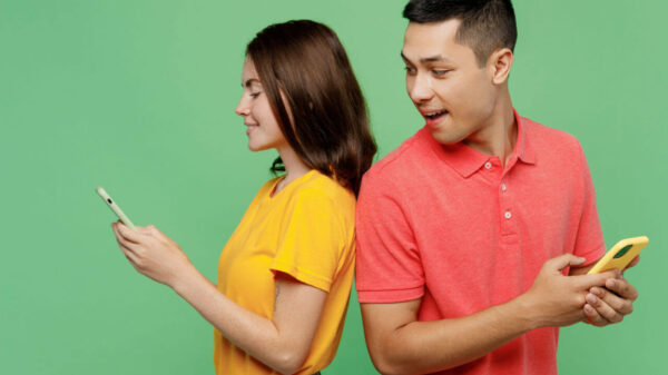 Casal de amigos jovens, (homem e mulher) usando camisetas básicas, juntos, cada um segurando um celular nas mãos, usando-os. Homem espiando o seu celular da mulher. De costas um para o outro, isolados em um fundo de cor verde pastel.
