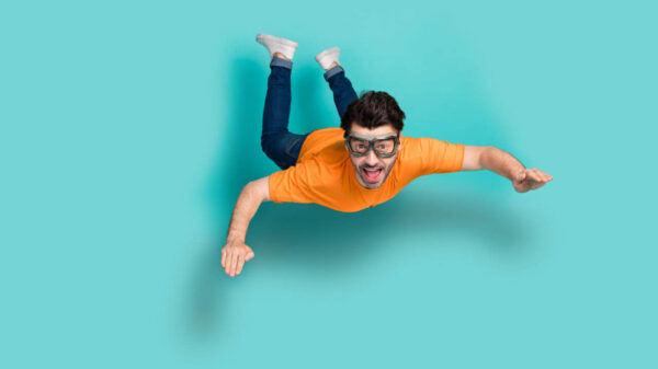 Foto do corpo inteiro de um jovem animado e positivo, usando óculos, saltando de um pára-quedas, pára-quedista, em queda livre, isolado em fundo de cor água-marinha.