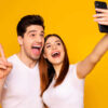 Retrato de homem e mulher alegres, positivos tirando selfie. Homem fazendo sinal de "v" com a mão. Isolados sobre um fundo amarelo.