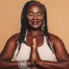 mulher negra de tranças com com as mãos juntas e roupas de ioga em fundo marrom