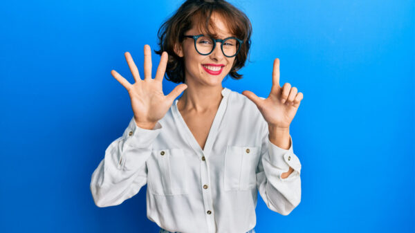 Mulher morena vestindo roupas casuais e usando óculos, fazendo com os dedos o número sete, enquanto sorri confiante e feliz.