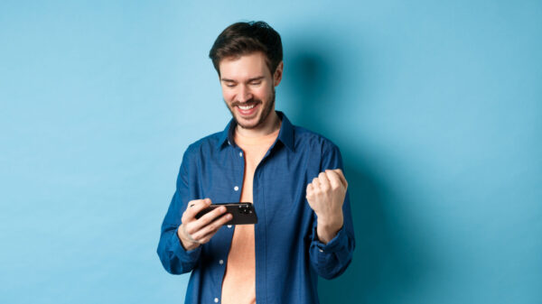 homem com expressão de felicidade segurando um celular na mão esquerda em fundo azul