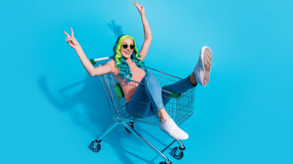 Retrato de uma mulher de bom humor, divirtindo-se, sentada dentro de um carrinho de compras, fazendo o sinal de "v" com as duas mãos, isolada em um fundo de cor azul.