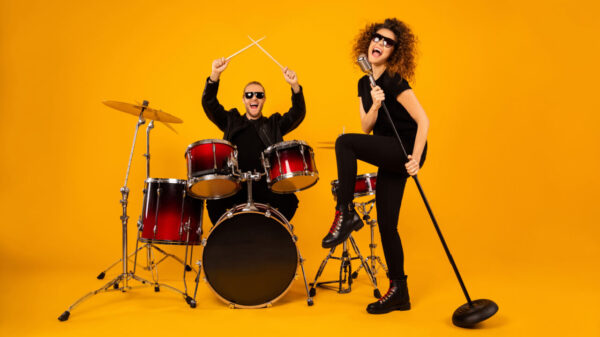 Foto de corpo inteiro de um grupo de rock. Homem tocando bateria e mulher cantando em um microfone. Conceito de banda popular. Os dois vestem roupas pretas modernas, e estão isolados em um fundo amarelo.