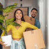 casal heterossexual entrando em casa carregando caixas e um vaso de planta