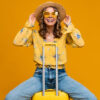Mulher loira sorridente, feliz, viajando com uma mala, vestida com roupas modernas, usando blusa, chapéu e óculos de sol, em um fundo amarelo, Conceito de férias.