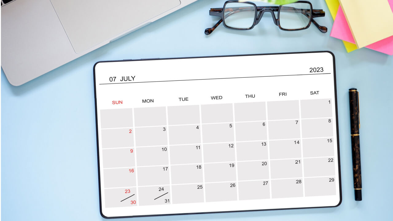 Vista superior de um notebook, de um tablet mostrando o calendário de julho de 2023, de uma caneta e de um óculos, em um fundo azul.