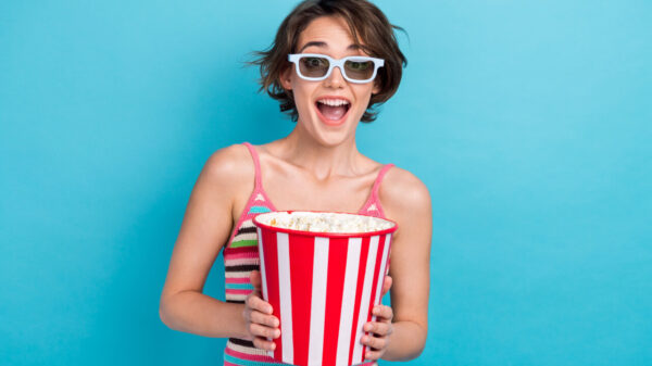 Foto de uma mulher alegre, chocada e surpresa, de boca aberta, segurando um balde com pipocas, usando um óculos para filmes 3D, isolada em um fundo de cor azul.