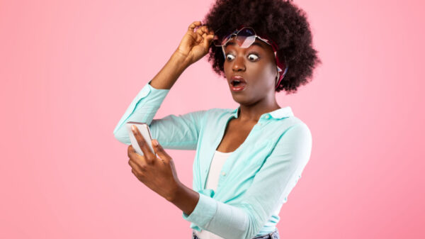 Mulher extremamente chocada, olhando para smartphone, levantando seus óculos com uma mão, posando sobre um fundo rosa de um estúdio.