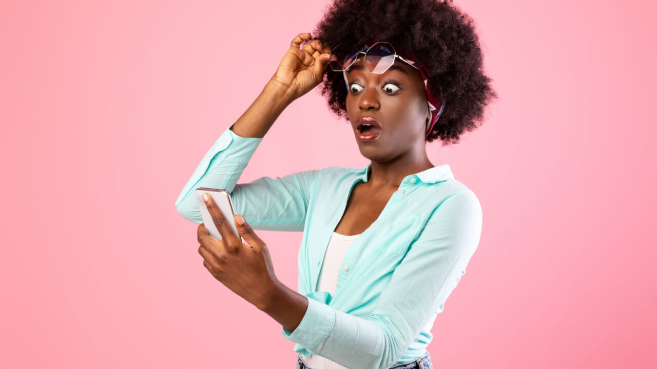 Mulher extremamente chocada, olhando para smartphone, levantando seus óculos com uma mão, posando sobre um fundo rosa de um estúdio.