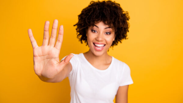 Foto de mulher fazendo o número 5 com os dedos de uma mão, vestindo uma camiseta branca casual, isolada, em um fundo amarelo.