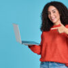 Mulher jovem, feliz, apontando para um laptop, isolada em fundo azul. Mulher sorridente segurando um computador.