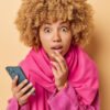 Retrato de uma mulher de cabelo encaracolado espantada, com a boca aberta, maravilhada, segurando um celular, vestido roupas rosas, isolada sobre um fundo bege. Conceito de reações humanas.