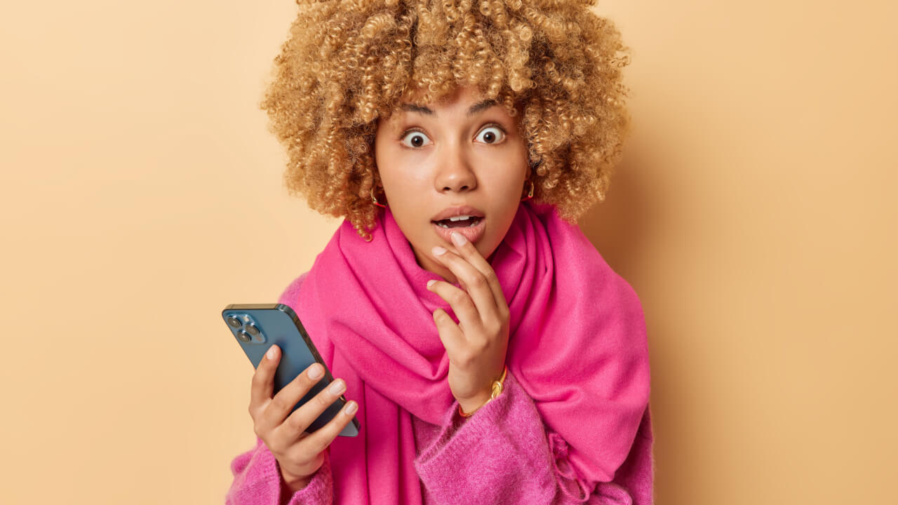 Retrato de uma mulher de cabelo encaracolado espantada, com a boca aberta, maravilhada, segurando um celular, vestido roupas rosas, isolada sobre um fundo bege. Conceito de reações humanas.